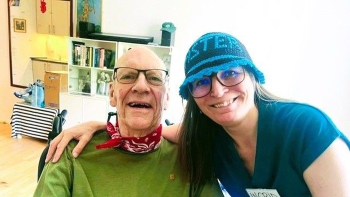 To personer smiler varmt, hvor den ene har en grøn skjorte og et rødt halstørklæde på, og de omfavner hinanden i en hyggelig indendørs indstilling.