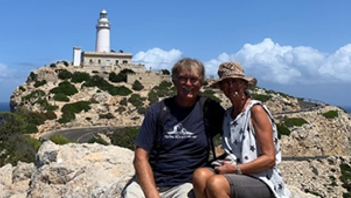 To personer poser sammen på et klippefyldt terræn med et fyrtårn i baggrunden under en klar himmel.
