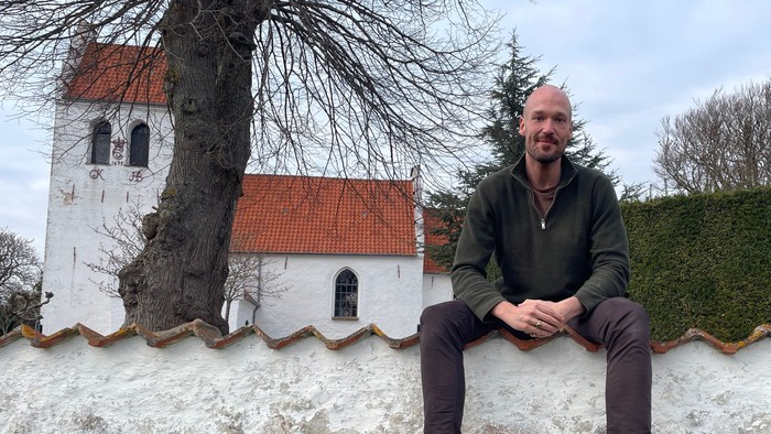En mand sidder på en stenvæg ved siden af en idyllisk kirke med rødt teglstenstag og nøgne træer.