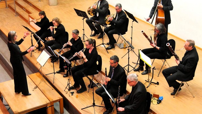 Ein Orchester tritt in einem geräumigen Saal auf, wobei Musiker verschiedene Instrumente unter der Leitung eines Dirigenten spielen.