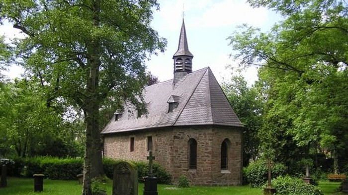 Kleine Steinkirche mit Turm, umgeben von Bäumen und Grabsteinen.
