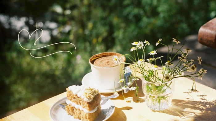 Ein Stück Kuchen und eine Tasse Kaffee auf einem Holztisch im Freien, begleitet von einer kleinen Vase mit Blumen