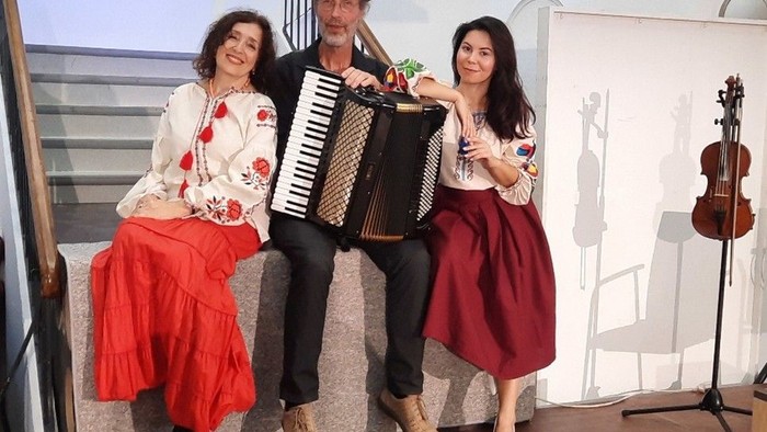 Drei Musiker posieren gemeinsam, wobei der Akkordeonspieler in der Mitte steht und Violinen zu sehen sind.