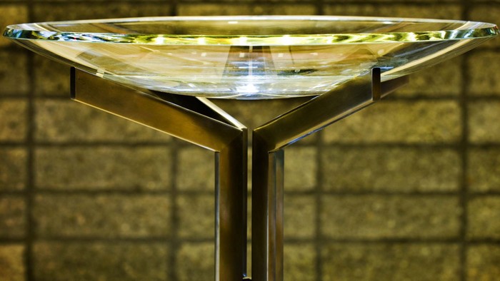 Moderner Cocktailtisch mit Glasplatte und schlankem Metallgestell vor strukturierter Wandkulisse.