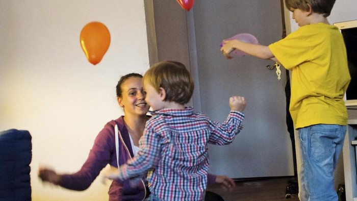 Eine Frau und zwei Kinder spielen fröhlich drinnen mit bunten Luftballons und einer Wii-Fernbedienung.