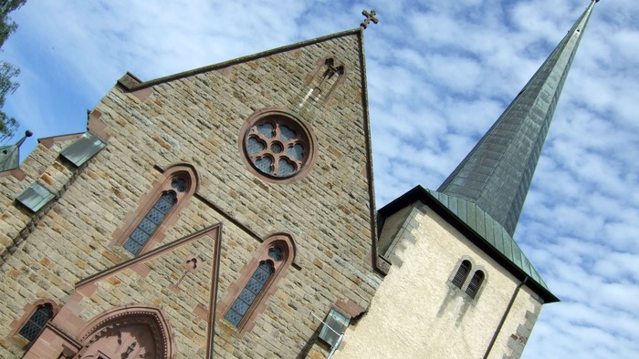Historische Steinkirche mit spitzem Turm und rundem Rosenfenster vor blauem Himmel.