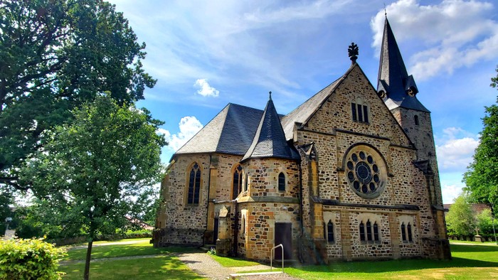Altes steinernes Kirchgebäude mit spitzen Türmen, umgeben von Bäumen und Gras.