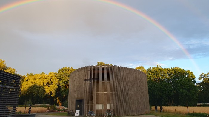 Ein beeindruckender Regenbogen wölbt sich über ein rustikales Holzgebäude, umgeben von üppigem Grün und abgestellten Fahrrädern.