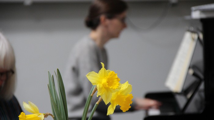 Forrest ser man en lille buket påskeliljer, og bagved (sløret) en kvinde, der spiller på klaver