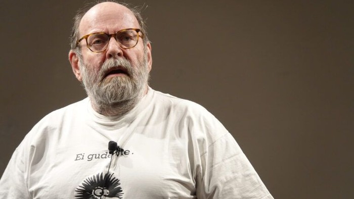 Ein älterer Mann mit Brille und grauem Bart steht vor neutralem Hintergrund und blickt nach vorne. Er trägt ein helles T-Shirt mit aufgedruckter Zeichnung und Schriftzug sowie ein Ansteckmikrofon, was auf einen Vortrag oder eine Bühnensituation hindeutet