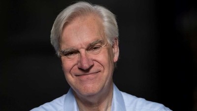 Mand med gråt hår, der smiler, iført en lyseblå knapskjorte på en mørk baggrund.