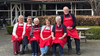 En gruppe på seks personer iført røde forklæder, der poserer sammen udendørs nær en rustik bygning.