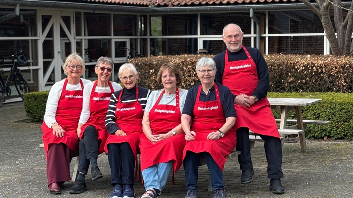 En gruppe på seks personer iført røde forklæder, der poserer sammen udendørs nær en rustik bygning.