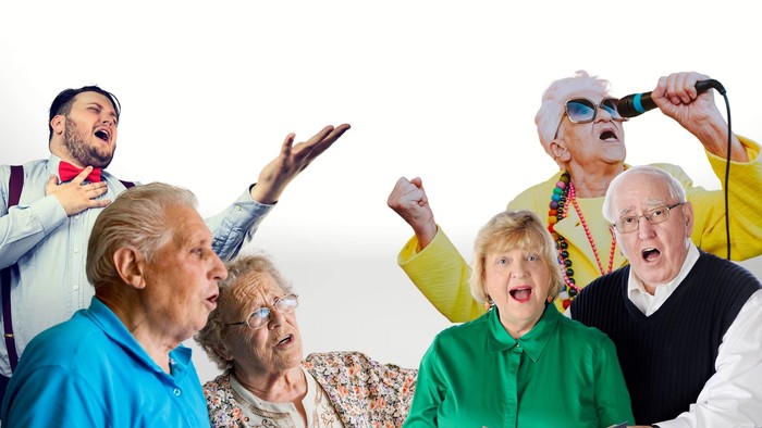 Eine Gruppe älterer Menschen feiert ausgelassen, einer hält ein Mikrofon, eine Frau liest aus einem Buch vor.