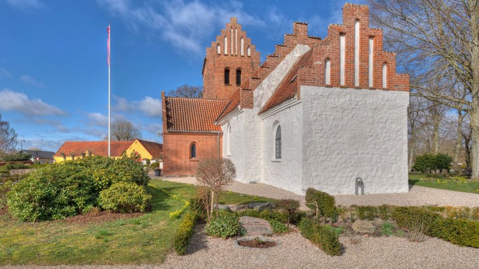 En lille kirke med røde mursten og hvidt tage, omgivet af en have og træer
