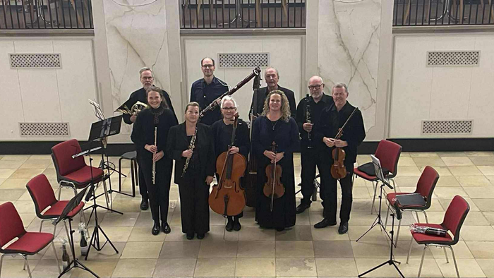 En gruppe musikere poser sammen med strenge- og blæseinstrumenter i en stor, elegant indendørs ramme.