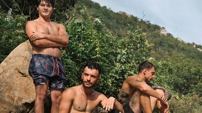 Drei oberkörperfreie Männer posieren im Freien in der Nähe von üppigem Grün und felsigem Gelände.