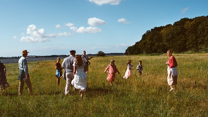 Eine Gruppe von Menschen in historischen Gewändern spaziert über eine grüne Wiese unter einem strahlend blauen Himmel.