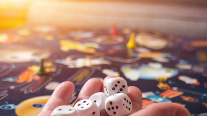 En hånd holder fem hvide terninger over et brettespil med farvede felter og figurer.