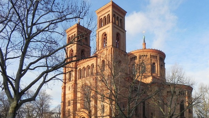 Historische Backsteinkirche mit Zwillingstürmen und rundem Chor, umgeben von kahlen Bäumen und parkenden Autos.