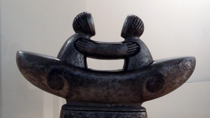 Abstrakte Steinskulptur, die zwei sich umarmende Figuren auf einem strukturierten Sockel darstellt.