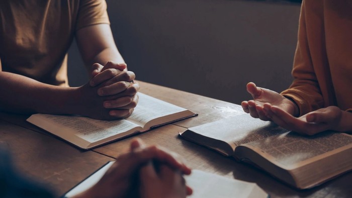 To personer sidder ved bord og læser bøger, en af dem har hænderne samlet.