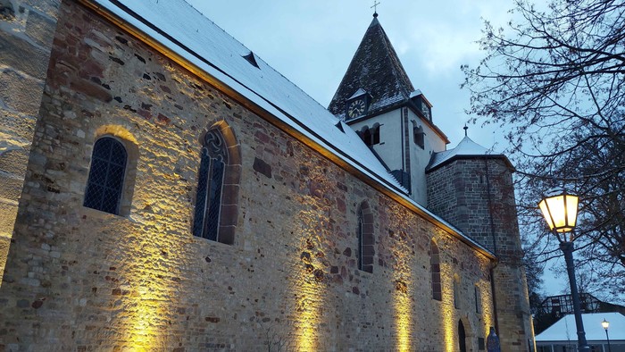 Historische Steinkirche nachts beleuchtet, mit leichtem Schnee bedeckt und einer Bank in der Nähe.