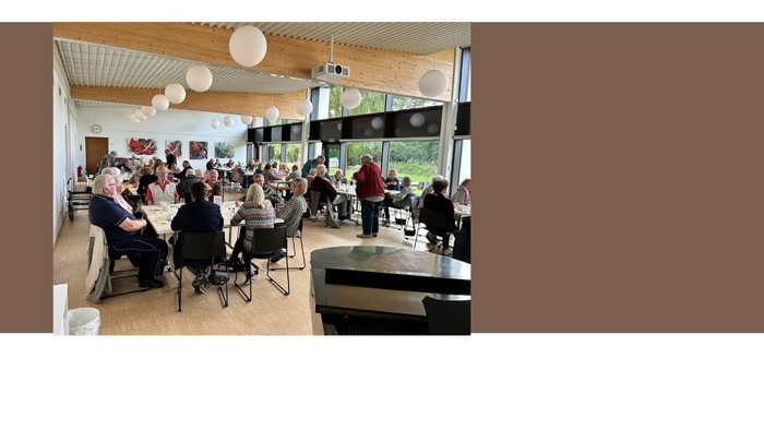 En gruppe af mennesker sidder i en moderne indendørs spiseområde med store vinduer og hængende lygter.