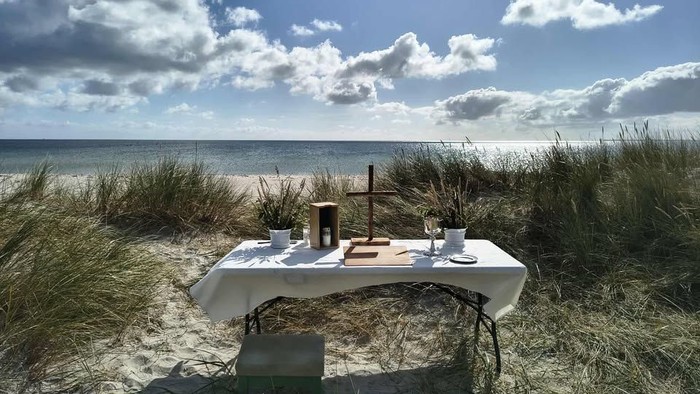 En rolig strandborddækning med enkle genstande og et kors med udsigt over havet under en delvist overskyet himmel.