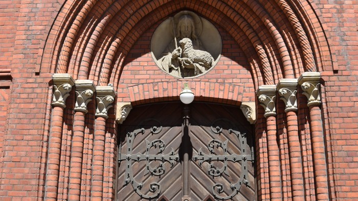 Die aus Backstein bestehende mit drei Säulen umrahmte Pforte der Herz Jesu Kirche. Im Triumphbogen ein Steinrelief von Christus, dem guten Hirten mit einem Lamm und einem Hirtenstab. Die Pforte aus dunklem Holz mit schmiedeisernen großflächigen Beschlägen