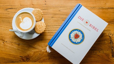 Ein Buch mit dem Titel "Die Bibel" liegt neben einer Tasse Kaffee und Keksen auf einem Holztisch