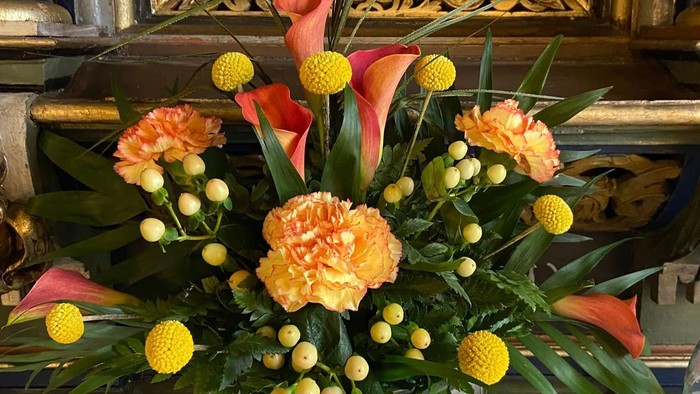 Livligt buket med orange nelliker, gule pomponblomster og grønt løv på et bord ved siden af et glas.