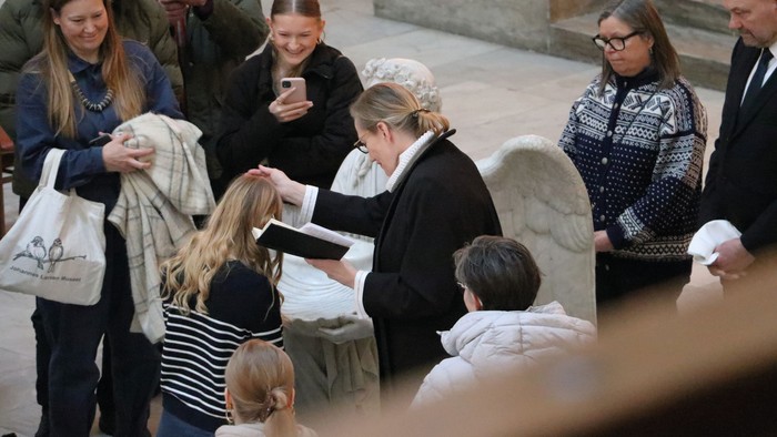 En gruppe af mennesker, heriblandt børn og voksne, er samlet om en grav. De holder blomster og blomsterkranser, og nogle af dem tager billeder.