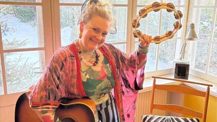 En person holder en guitar og en dekorativ brødring nær et hyggeligt, farverigt indendørs miljø med udsigt gennem et vindue.