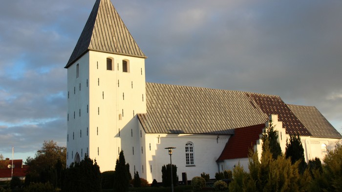 Nørre Løgum kirke, set fra syd/vest med grå skyer og lidt blå himmel som baggrund. Solen skinner. Kirken er hvidkalket, har et tårn og der er bly på taget. Forrest ses grønnehække og buske på kirkegården