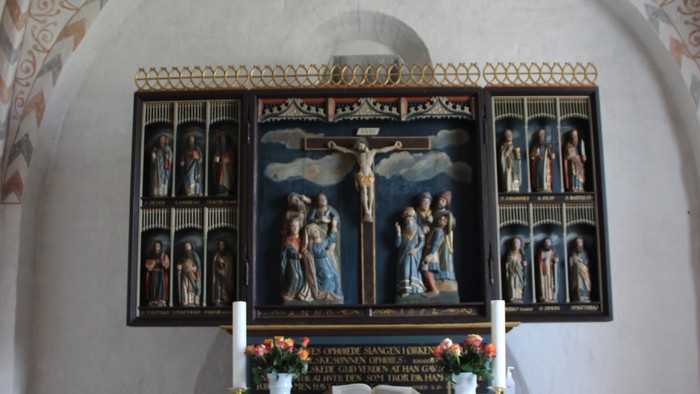 Alter Nr. Løgum kirke, hvorover der ses loftets buer med et kalkmaleri-mønster malet på i sarte farver. Alteret er beklædt med rødt stof og øverst ligger den hvide alterdug, som har en kniplet kant med kors på. Altertavlen er tre-fløjet. I midten ses Jesu