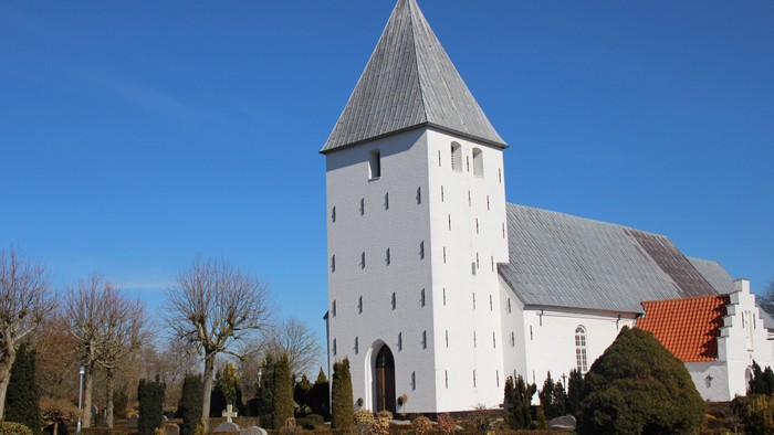 Nr. Løgum kirke og kirkegård fra sydvest med blå himmel som baggrund. Solen skinner og lindetræerne står nøgne med deres korte grene.