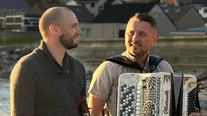 To musikere, den ene spiller violin og den anden harmonika, står ved en havnefront under solnedgang.