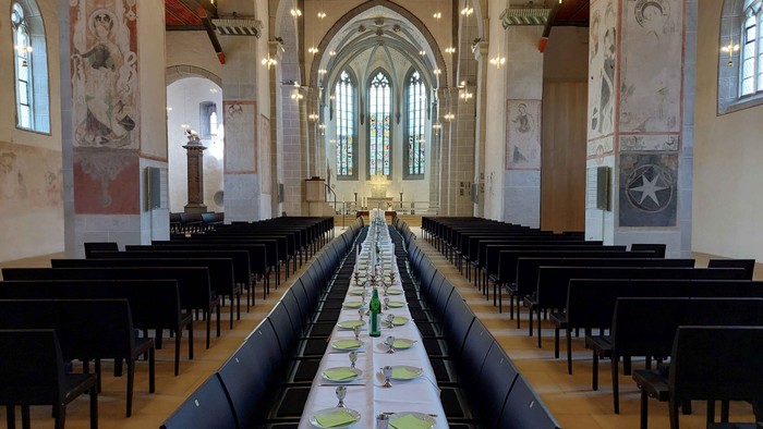 Großer Esstisch gedeckt mit Tellern und Gläsern in einem geräumigen Saal mit hohen Decken und Rundbogenfenstern.