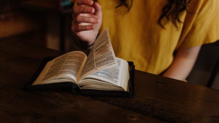 Eine Person liest ein aufgeschlagenes Buch mit gefalteten Händen über den Seiten auf einem Holztisch.