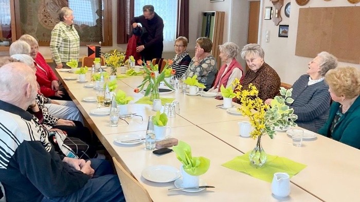 Gruppe älterer Menschen, die an einem langen Tisch mit gedeckten Plätzen und Vasen mit Blumen sitzen.