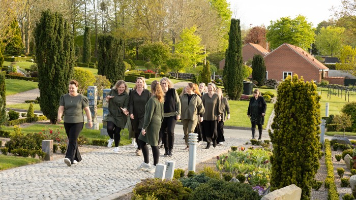 En gruppe mennesker går gennem en velholdt kirkegård på en solrig dag.