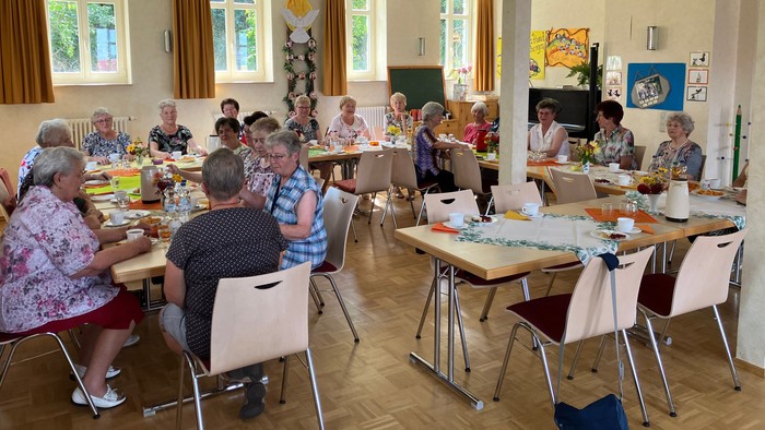 Eine Gruppe von Menschen sitzt an Tischen und isst gemeinsam in einem hellen Raum mit großen Fenstern.
