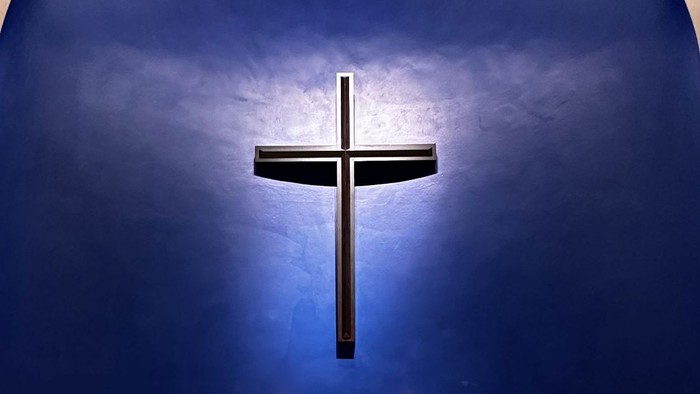 Ein weißer Kerzenleuchter steht vor einem großen Holzkreuz an einer blauen Kirchenwand