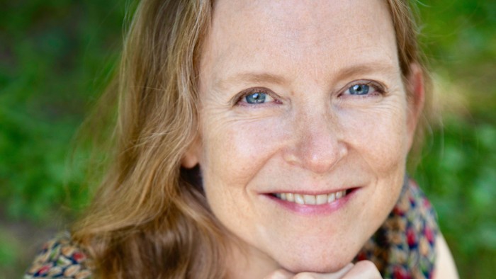 Kvinde med langt hår, der smiler blidt, mens hun hviler hagen på hånden udendørs.