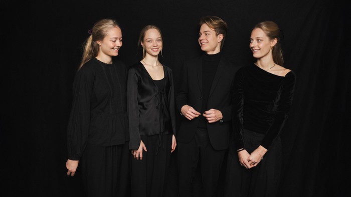 Fire personer i formelt sort tøj, der smiler sammen mod en mørk baggrund.