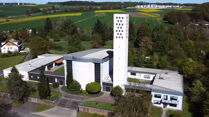 Moderner Kirchenkomplex, umgeben von üppigem Grün und landwirtschaftlichen Feldern mit Windkraftanlagen im Hintergrund.