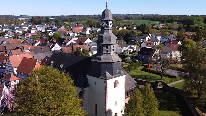 Luftaufnahme eines malerischen Dorfes mit einer markanten Kirche, umgeben von bunten Häusern und üppigem Grün.