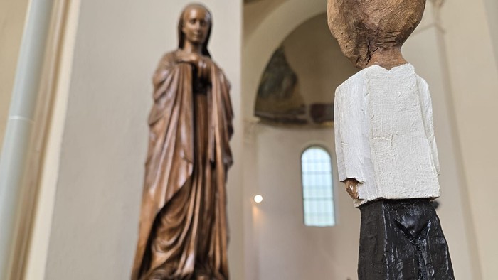 Die Statue eines Kindes mit großen Kopf steht vor einer Jesus-Statue in einer Kirche.