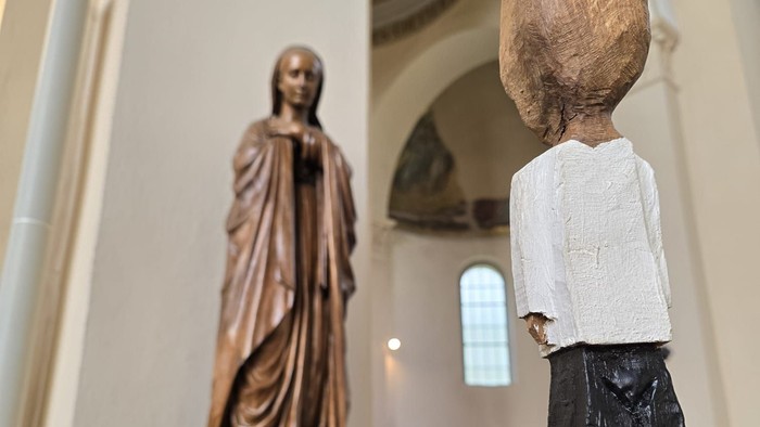 Die Statue eines Kindes mit großen Kopf steht vor einer Jesus-Statue in einer Kirche.